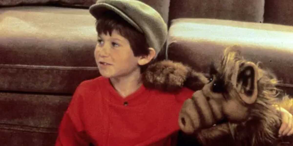A young boy wearing a grey flat cap and a red shirt sits on the floor, smiling. Next to him is an alien puppet with brown fur, large snout, and big ears, resting its arm on the boy's shoulder. Behind them is a brown sofa. This beloved child actor's joy shines through every scene they share.