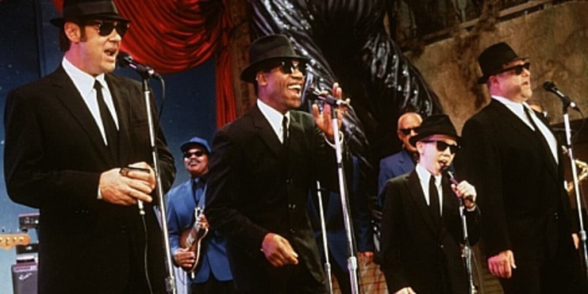 A band dressed in black suits, white shirts, thin black ties, and black sunglasses performs energetically on stage. Four members sing into microphones while a fifth member plays saxophone in the background. The stage is decorated with red and blue drapes.