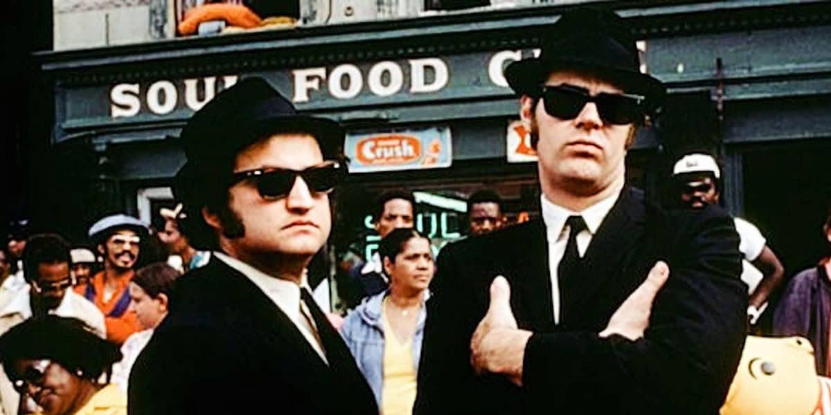 Two men are standing in front of a "Soul Food Cafe" wearing black suits, black hats, and dark sunglasses. One has his arms crossed, and the other stands with his arms at his sides. The background features a crowd of people and a "Crush" soda sign.