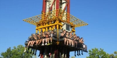 A group of people are seated on a thrill ride at a theme park, suspended in mid-air. They are secured by harnesses and appear to be anticipating a drop. The ride structure is red and yellow, and the background shows a clear blue sky and green trees.