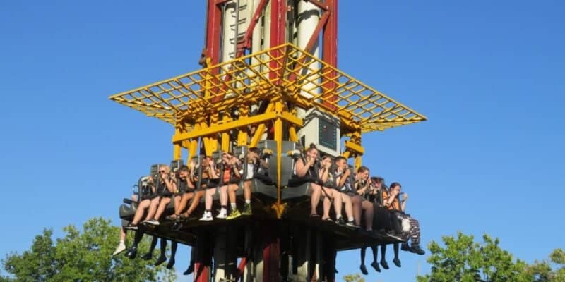 A group of people are seated on a thrill ride at a theme park, suspended in mid-air. They are secured by harnesses and appear to be anticipating a drop. The ride structure is red and yellow, and the background shows a clear blue sky and green trees.