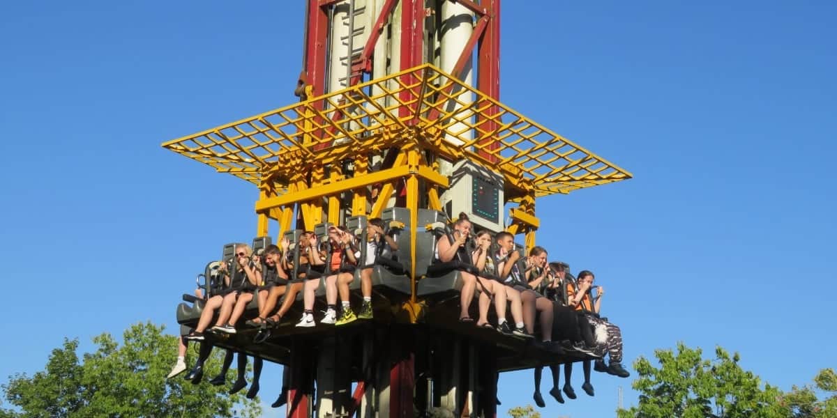 A group of people are seated on a thrill ride at a theme park, suspended in mid-air. They are secured by harnesses and appear to be anticipating a drop. The ride structure is red and yellow, and the background shows a clear blue sky and green trees.
