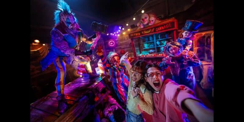 A group of people are screaming in delight as they pose for a selfie at Halloween Horror Nights in a haunted house. The setting is filled with eerie lights, creepy dolls, and costumed performers in suspenseful clown and circus-themed attire, intensifying the spooky atmosphere.