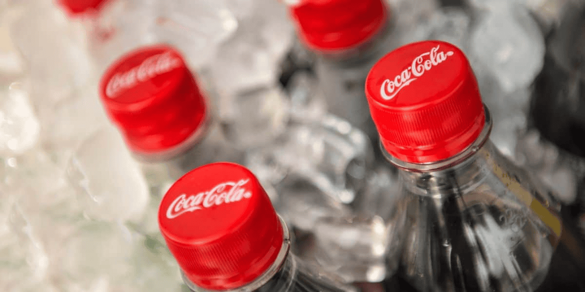 Close-up of several Coca-Cola bottles with red caps, partially submerged in ice. The focus is on the caps, each labeled with the Coca-Cola logo. Perfect for quenching thirsts, whether you're entertaining guests or soaking up the magic at Disney World. The ice cubes around the bottles appear partially melted.