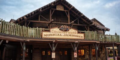 A rustic log cabin-style building with a sign that reads "Country Bear Musical Jamboree" hanging above the entrance. The building has a wooden fence on the balcony, lantern-style lights, and a cloudy sky in the background.