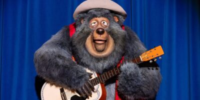 A large bear character with thick, dark fur plays an acoustic guitar. The bear is wearing a gray hat and a red vest and is positioned in front of a blue curtain, giving the appearance of performing on stage.