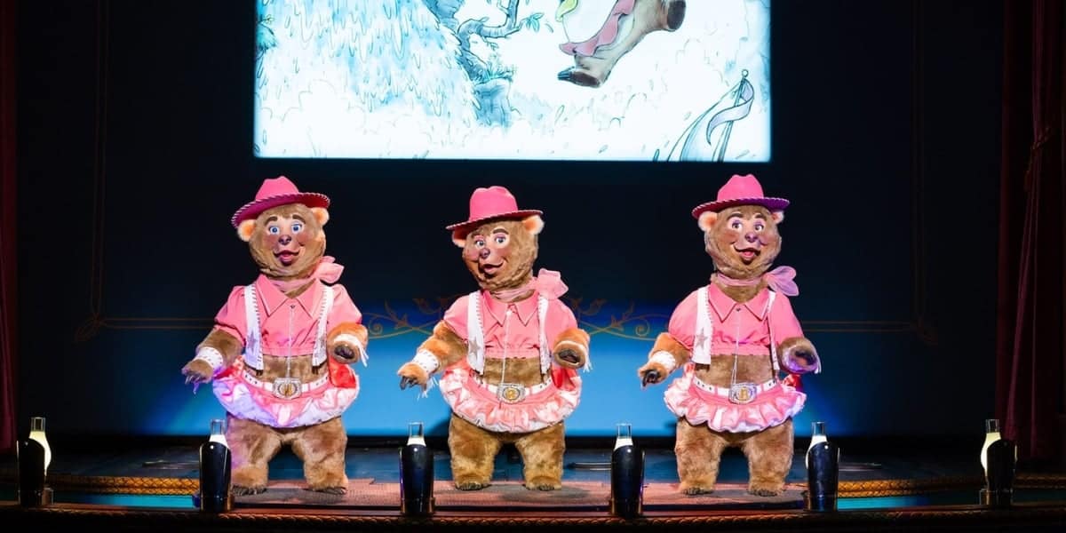Three anthropomorphic bears wearing pink cowboy outfits and hats are dancing on a stage. Behind them is a screen with a blue and white illustration. They are positioned in front of several black bottles lined up on the floor.