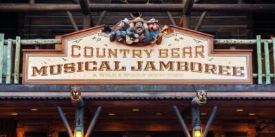 A wooden sign for "Country Bear Musical Jamboree" is displayed, featuring three animated bear characters in hats above the text. The subtitle reads "A Wild & Wooly Good Time." The sign is part of a rustic, log cabin-styled building with balcony rails.
