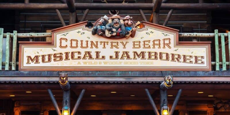 A wooden sign for "Country Bear Musical Jamboree" is displayed, featuring three animated bear characters in hats above the text. The subtitle reads "A Wild & Wooly Good Time." The sign is part of a rustic, log cabin-styled building with balcony rails.