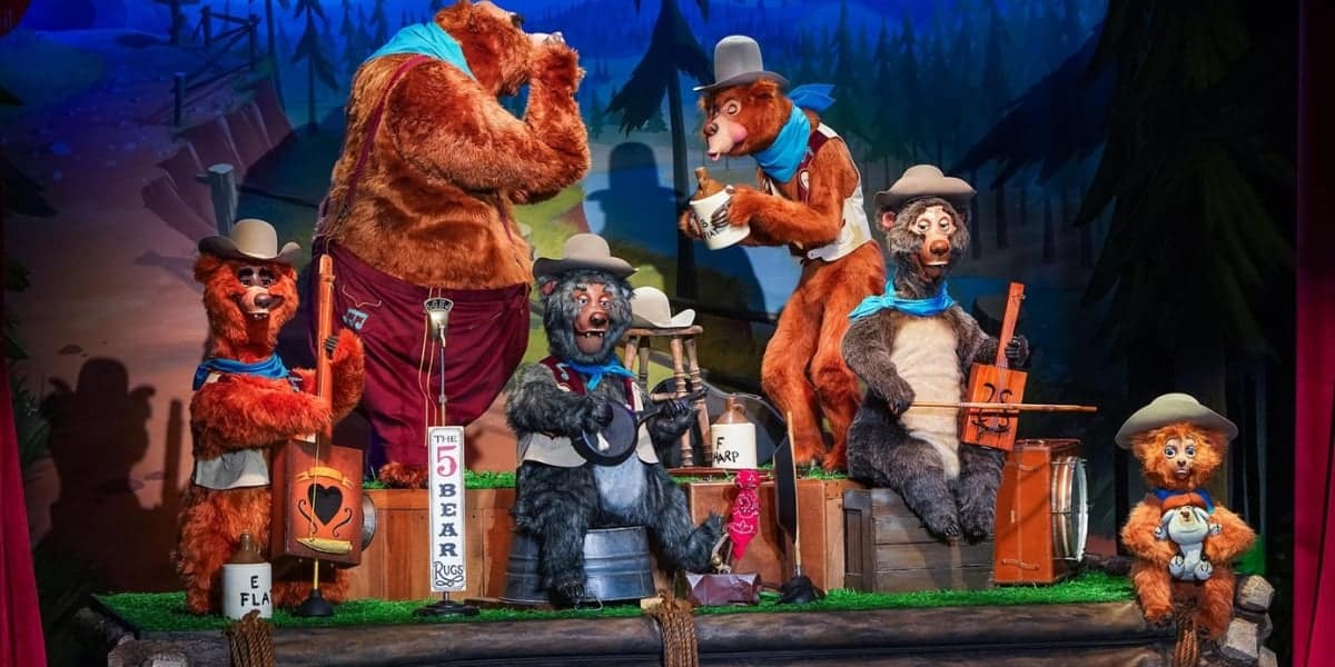 A group of animatronic bears wearing hats and scarves perform music on stage. They play various instruments, including a banjo, a guitar, and a fiddle. The background features a forest scene, and the foremost bear is dressed in red pants and a blue shirt.