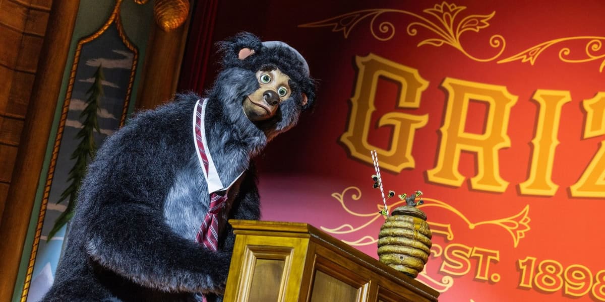 An animatronic bear wearing a red striped tie stands behind a wooden podium. In front of it is a jar shaped like a beehive with utensils in it. The backdrop has vintage-style red and yellow lettering, reading "GRIZ" with some decorative designs.