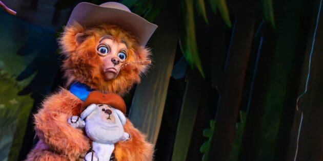 A brown, furry bear character in a cowboy hat and blue scarf holds a plush bear wearing a red hat. The background features a dark, forest-like scene with green foliage and trees. The bear has a surprised expression.