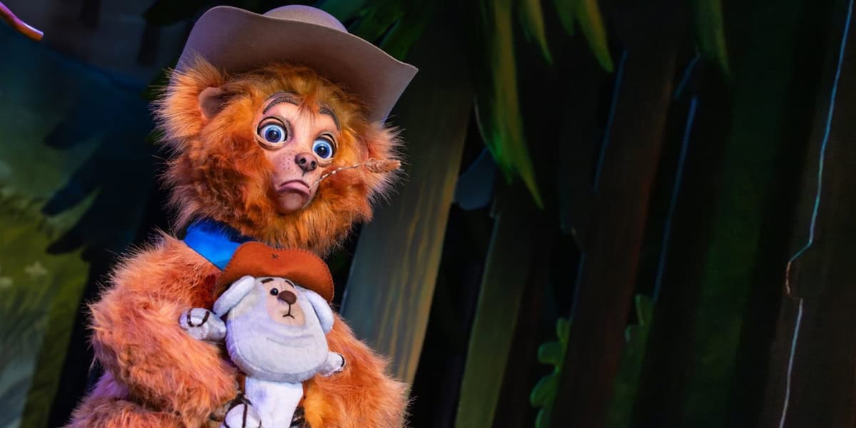 A brown, furry bear character in a cowboy hat and blue scarf holds a plush bear wearing a red hat. The background features a dark, forest-like scene with green foliage and trees. The bear has a surprised expression.