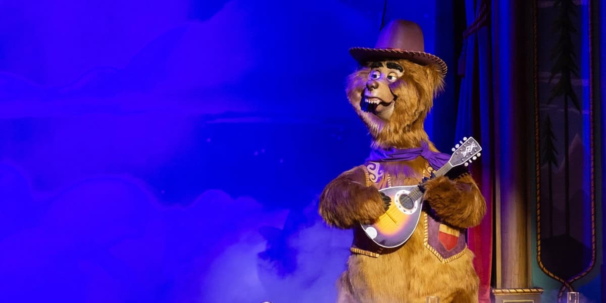 A brown bear in a western outfit, including a cowboy hat and bandana, stands on stage. The bear is playing a small stringed instrument, possibly a mandolin or ukulele. The background features purple mist and a colorful backdrop, suggesting a performance setting.