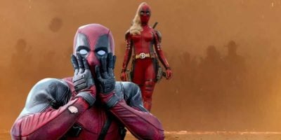 A character in a red and black costume, resembling Deadpool, is in the foreground holding their face in a surprised gesture. In the background, another character in a similar costume with long blonde hair stands confidently against a smoky orange backdrop.