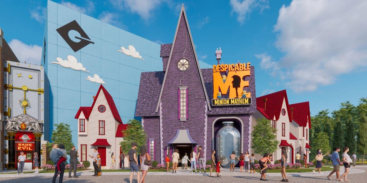 A colorful theme park scene at Universal Studios features the "Despicable Me Minion Mayhem" attraction. Visitors walk around the whimsical setup, which includes a purple house with a pointed roof, vibrant signs, and various Minion-themed decorations. The sky is clear and sunny.