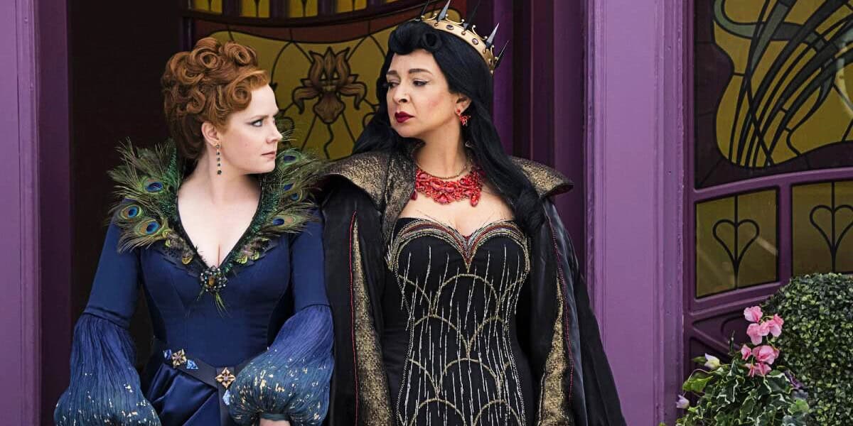 Two women dressed in elaborate costumes stand side by side. The woman on the left wears a blue, peacock feather-themed gown, while the woman on the right wears a black and gold gown with a crown. They stand in front of a purple door and arched windows with stained glass.