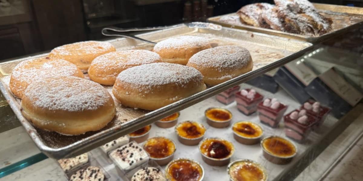 A display case filled with assorted pastries and desserts. The foreground features powdered sugar-covered donuts on a tray. Behind them are pastries dusted with powdered sugar. The bottom shelf displays various desserts, including crème brûlée and layered cakes.