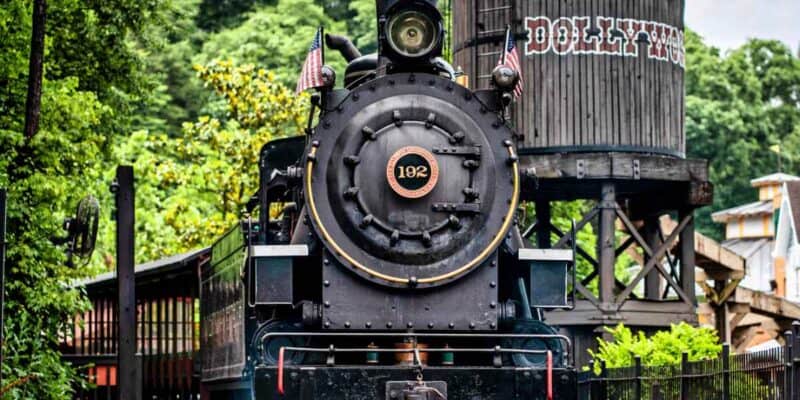 A vintage steam locomotive with a circular number plate "192" on its front stands on railway tracks surrounded by greenery. The background features a wooden water tower with "Dollywood" written on it, adding to the charm of Dolly Parton's famous theme park.