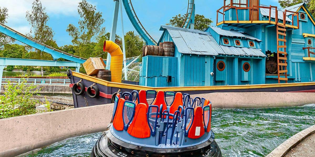 A circular raft for a white-water rapids ride is shown in the foreground. Behind it, there's a whimsical blue and orange themed ship adorned with barrels and small windows. The theme park setting is lush with trees, and looping roller coaster tracks are visible in the background.