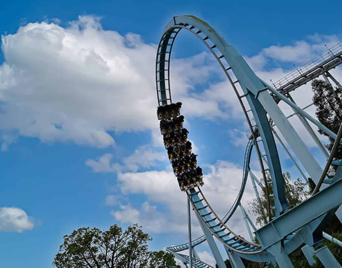 A large, blue steel roller coaster with a full vertical loop is set against a bright blue sky with a few white clouds. The coaster has a train of riders in the loop, partially upside-down. Theme park trees are visible in the lower part of the frame.