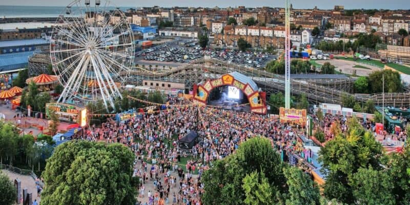 A large outdoor music festival with a massive crowd gathered in front of a stage. An amusement park, particularly popular for its Ferris wheel and roller coaster, looms in the background, surrounded by trees and buildings. The image is taken from an elevated vantage point.