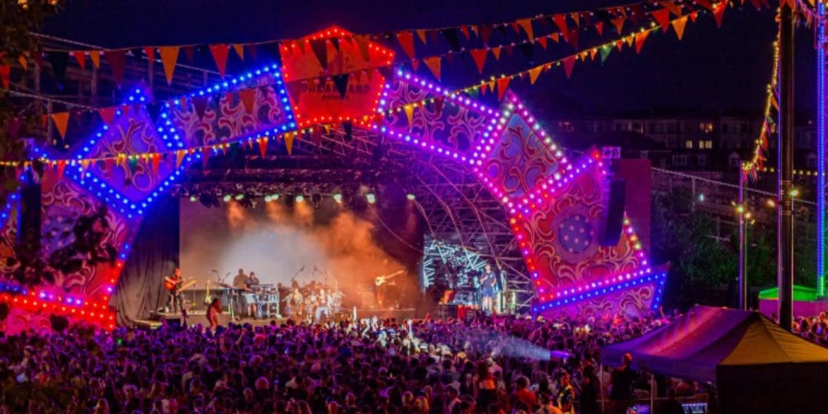 A lively outdoor concert scene at night with a large crowd in front of a brightly lit stage adorned with colorful decorations and illuminated patterns. Bunting and banners hang overhead, adding to the festive atmosphere of this popular event. Musicians are performing on stage.