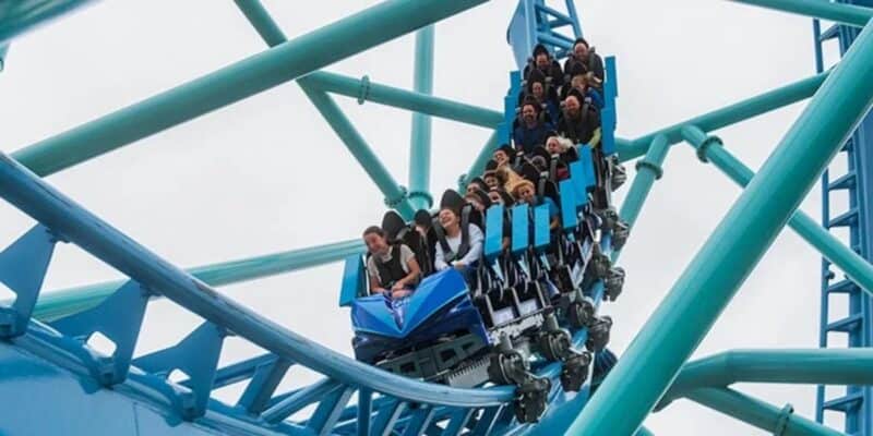 A group of people, seated in rows, ride a blue roller coaster at SeaWorld as it twists on a track with teal supports. The riders appear excited and animated despite the overcast sky watching over them at an Orlando theme park.