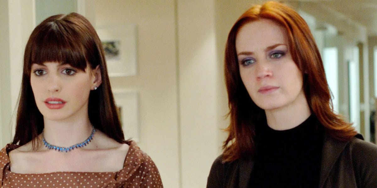 Two women stand indoors, facing forward. The woman on the left has long, straight brown hair with bangs, and is wearing a brown polka dot top and a blue necklace. The woman on the right has shoulder-length red hair and is wearing a black top with a brown jacket.