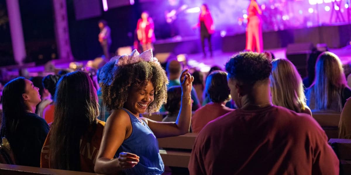 A group of people enjoys a live music performance. The audience is engaged, with one woman in the foreground dancing and smiling. The stage is lit with colorful lights, and musicians can be seen performing in the background.