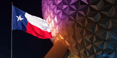 The image shows the Texas state flag waving on a pole with a background of a large geodesic sphere, lit up with various colors. The flag, representing the Lone Star State, consists of a blue vertical stripe with a white star and horizontal white and red stripes—welcoming guests like Disney World.