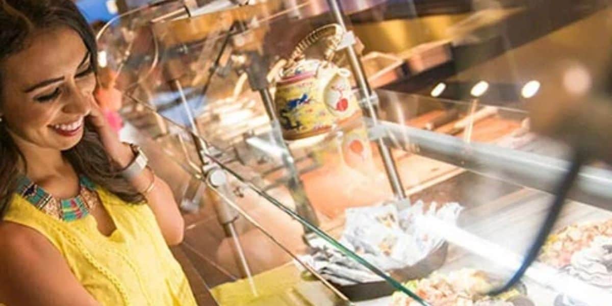 A woman in a yellow top with a colorful necklace looks at food in a buffet service line. She smiles and holds her hand to her face. The background features a teapot with dragon designs on a glass shelf.
