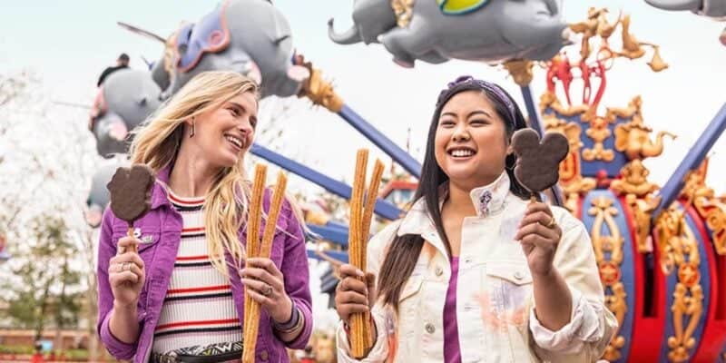 Two women are smiling and enjoying snacks at an amusement park. One holds two churros and a Mickey Mouse-shaped ice cream bar, while the other has an ice cream bar. Colorful flying elephant rides are visible in the background.