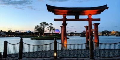 A traditional Japanese torii gate stands prominently near the edge of a lake at twilight. In the background, trees and a lit geodesic dome structure evoke scenes from Walt Disney World. The sky transitions from blue to orange as the sun sets, casting reflections on the water.