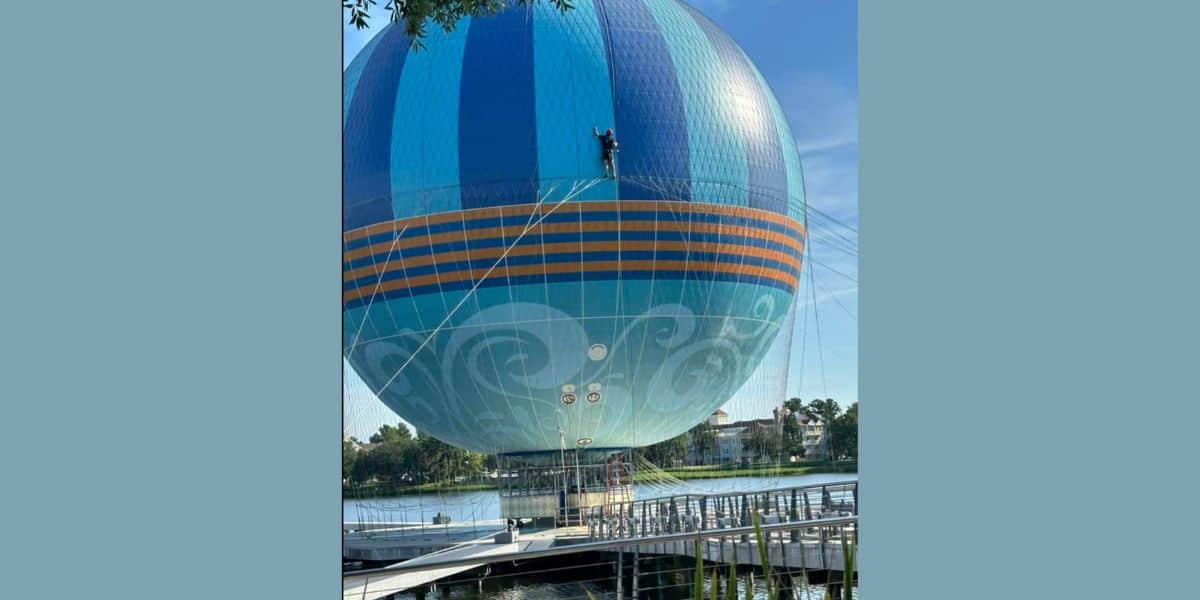 A large, blue and teal hot air balloon with orange stripes is tethered to the ground near a lakefront dock at Disney Springs. Swirling patterns are visible on the balloon as trees and buildings form the background under a clear sky.