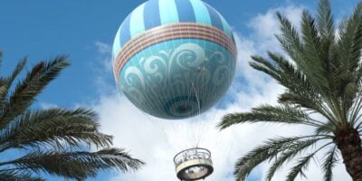 A blue hot air balloon with intricate swirled patterns and orange stripes ascends into a blue sky with fluffy white clouds above Disney Springs. The basket is visible below, and tall palm trees frame the scene on either side.
