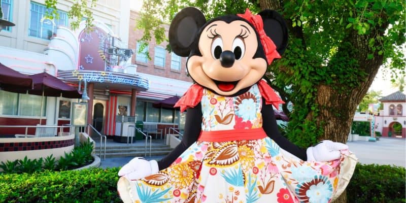 A costumed character, Minnie Mouse, stands outside under a tree. Minnie is wearing a vibrant floral dress and a red hair bow, smiling widely with both hands holding up the sides of her dress. The background displays an art deco-style building and restaurant.