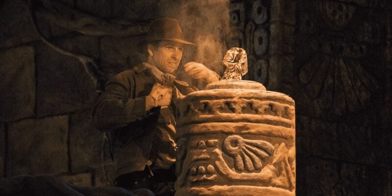 A person, dressed in adventurer gear including a fedora and leather jacket, carefully reaches towards a golden idol placed atop a stone pedestal in a dimly lit, ancient-looking setting with carved walls and dusty atmosphere.