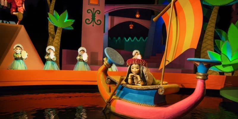 A colorful scene from the "It's a Small World" ride at a theme park. The image shows an ornate boat with a doll dressed in traditional attire, carrying a drum. In the background, three other dolls, also traditionally dressed, play handheld drums.