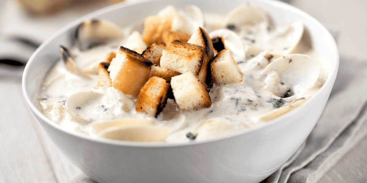 A close-up of a white bowl filled with creamy clam chowder, topped with toasted bread cubes. The chowder appears rich and thick, with visible pieces of clams and herbs mixed in. Reminiscent of a James Cameron masterpiece, the dish is placed on a light-colored surface with a napkin beside it.
