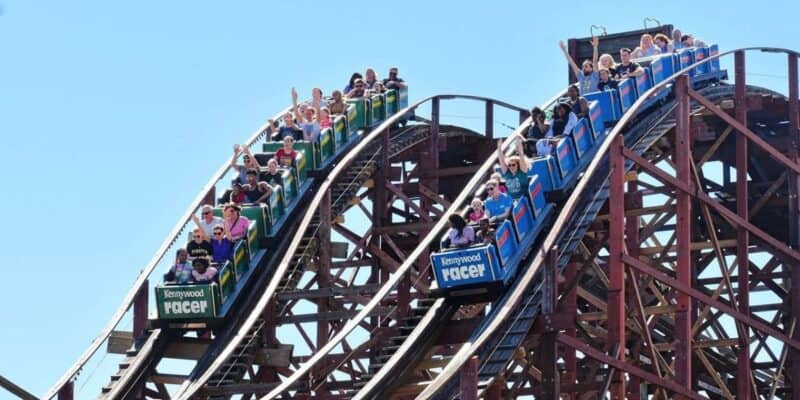 People riding the twin tracks of the 97-year-old wooden roller coaster named "Racer" at Kennywood theme park. The roller coaster has blue and green cars filled with excited riders, their hands up in the air against a clear blue sky.