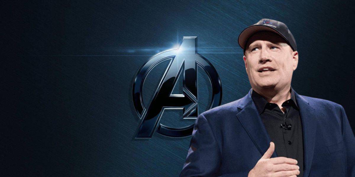 A man wearing a black cap and navy blue blazer speaks in front of a dark background featuring the Avengers logo, which is a stylized "A" with an arrow.