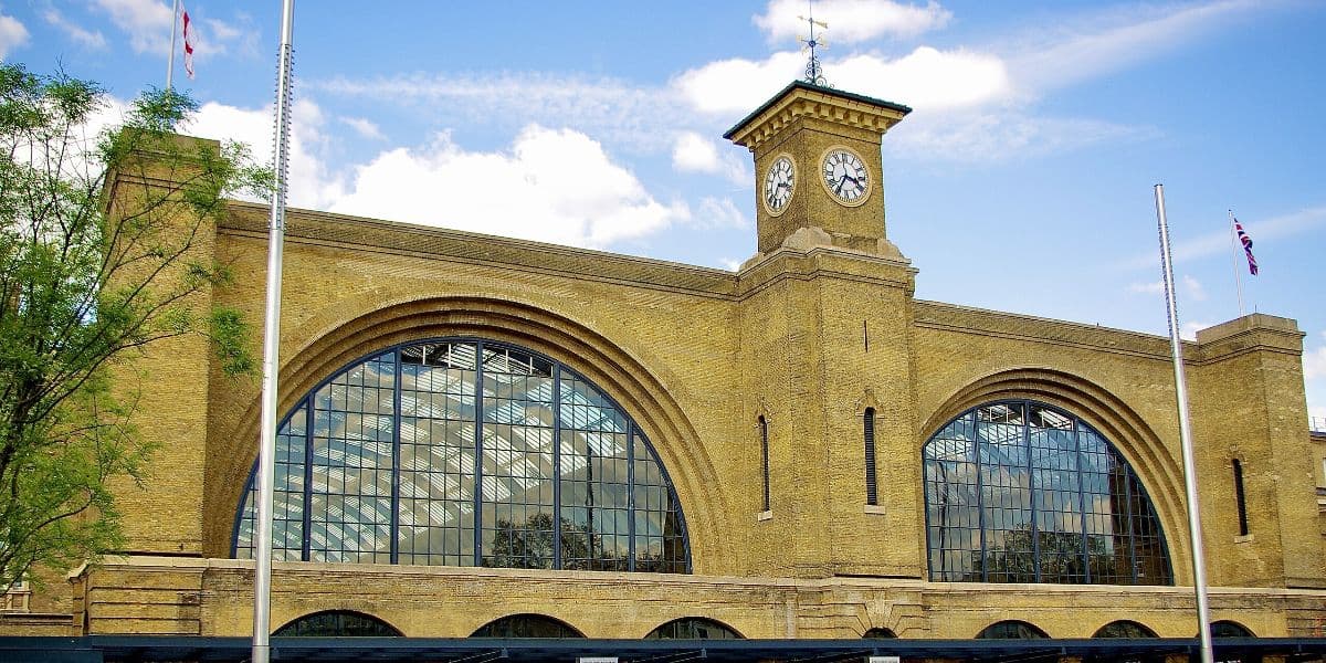 A large, historic railway station building with arched windows and a central clock tower. The structure is made of light brown brick and features intricate architectural details reminiscent of a magical attraction. Two flagpoles are visible on either side under a partly cloudy sky.
