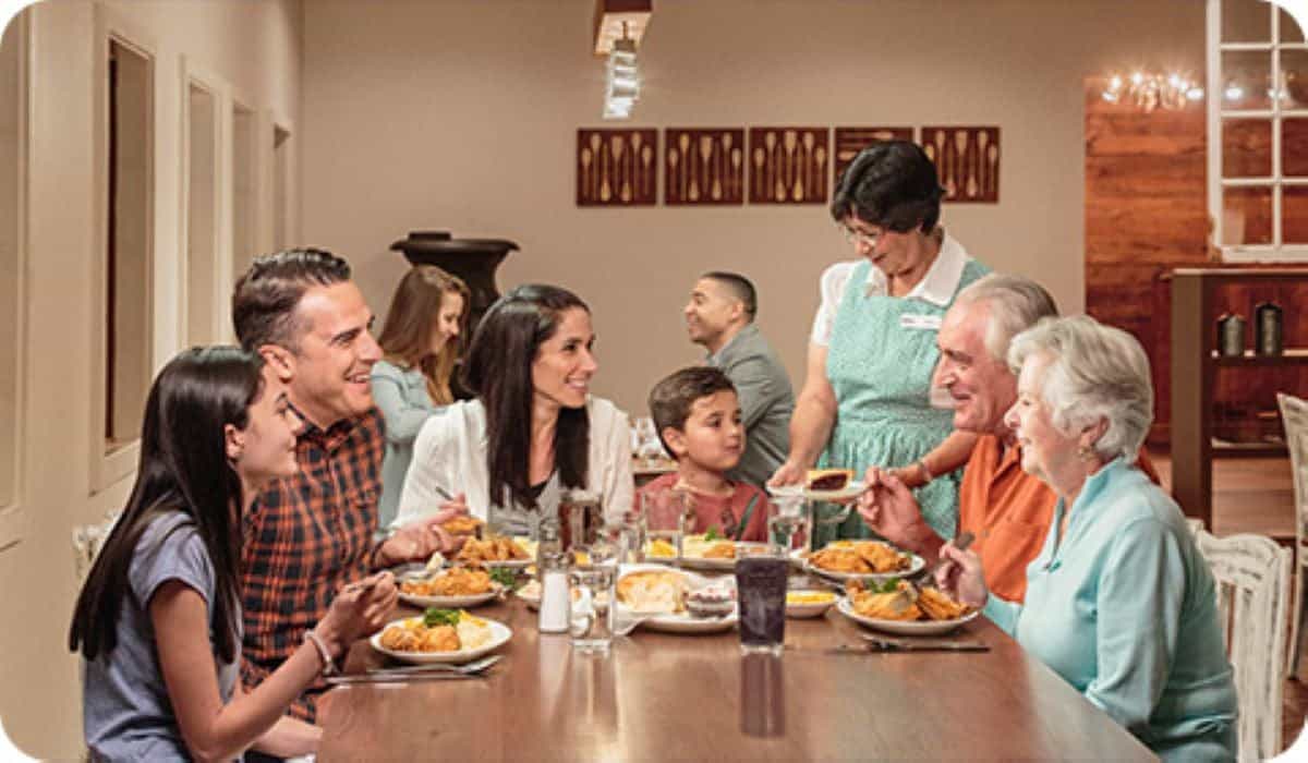 A diverse group of select guests sits around a long wooden table, enjoying a meal together. Two children and four adults converse and smile as a server in a green apron distributes food. The setting is warm and cozy, with brown and beige decor reminiscent of a California theme park.