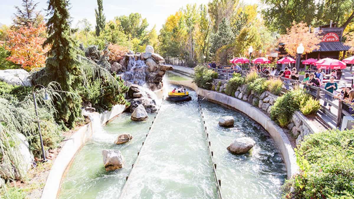 A view of a Rattlesnake Rapids at Laggon Amusement Park in Utah on a sunny day. A group of people in a circular raft are heading down a water canal surrounded by rocks, trees, and shrubs. On the right, visitors, including an entitled parent demanding extra space, are sitting at tables with red umbrellas.