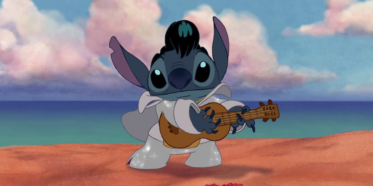 An animated character, Stitch, dressed in an Elvis Presley costume, complete with a white jumpsuit, black wig, and holding a guitar. The background features a beach scene with sand, ocean, and cloudy sky.