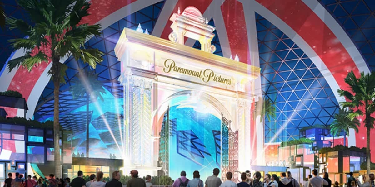 A vibrant nighttime scene at a theme park entrance. The illuminated, grand archway bears the "Paramount Pictures" logo. Colorful lights and projections adorn the surrounding structures, reminiscent of Disneyland's magic. People stand in the foreground, enjoying the lively atmosphere with palm trees on either side.