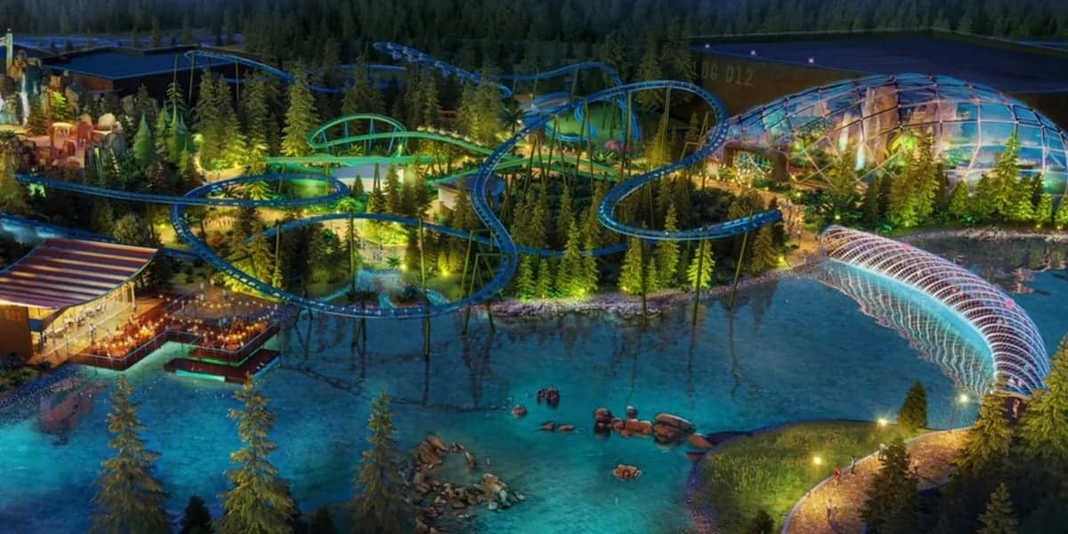 An illuminated amusement park at night, surrounded by dense woodland. The Theme Park Hopes feature a winding roller coaster, a glass-covered structure, a bridge with light displays over a river, and various lit-up buildings, creating a vibrant and magical atmosphere.