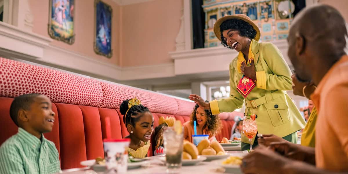 A joyful restaurant scene with a family seated in a booth. A woman in a bright outfit and hat stands next to the table, smiling and engaging with the children, one of whom is dressed as a princess. The family is surrounded by food and drinks on the table.