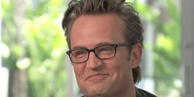 A middle-aged man with short, greying hair smiles softly. He is wearing black-rimmed glasses and a dark shirt. The background is blurred, showing some greenery and indoors lighting, reminiscent of the peaceful scenes in Friends where Matthew Perry's iconic character found solace.